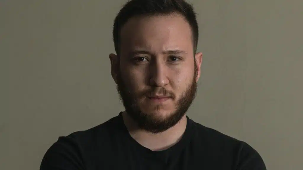 A close-up studio portrait of a bearded man in a black shirt looking directly at the camera.
