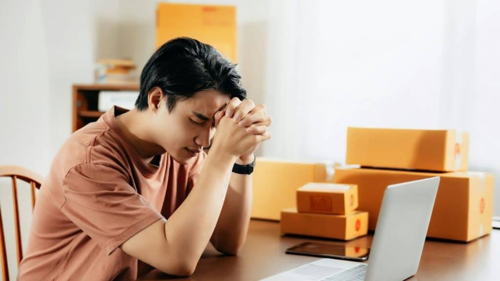 A stressed man sits at a desk with a laptop, his head bowed into his clasped hands.