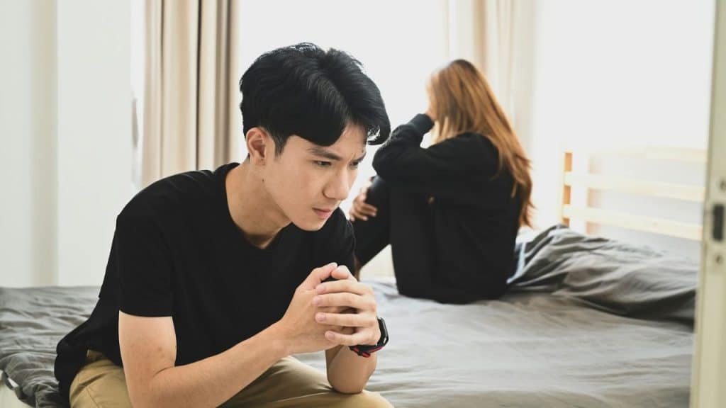 A man sits forlornly on a bed while a woman sits crying in the background.
