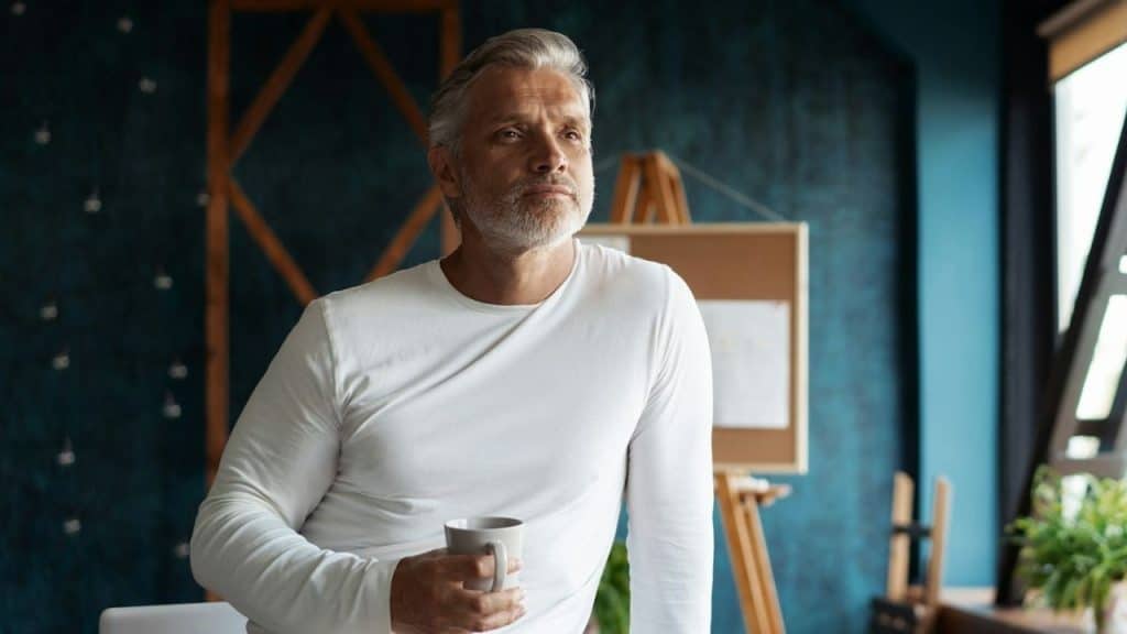 Gray-haired man in a white shirt holding a mug, looking into the distance.