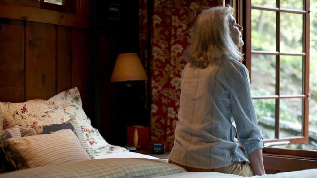 Elderly woman with gray hair sits on a bed, looking out an open window.