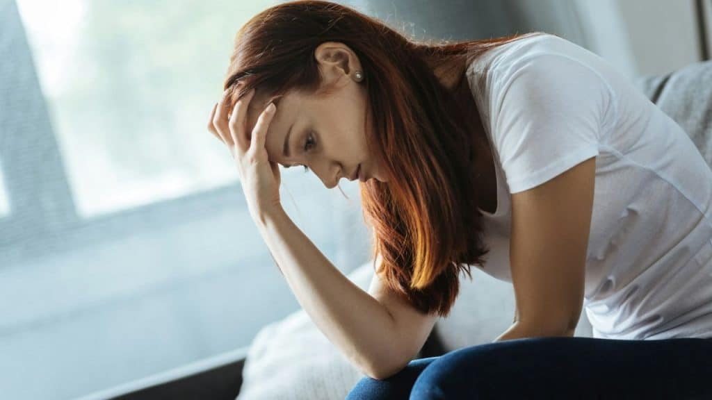 Red-haired woman in a white shirt sits slouched with her hand on her head.