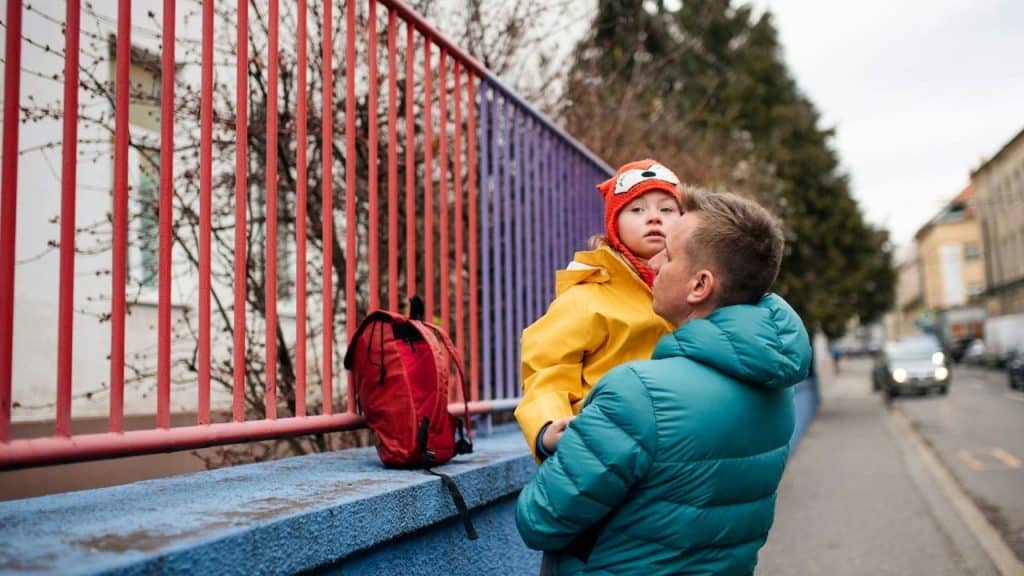 Father holding a child in a yellow coat next to a red and blue fence.