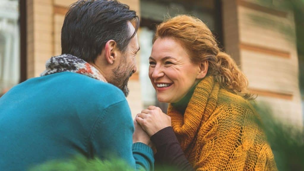 Middle-aged couple holding hands, smiling and gazing into each other's eyes outdoors.
