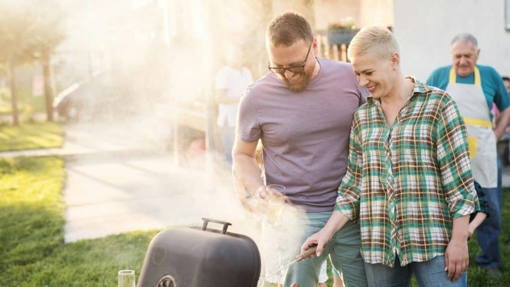 Smiling couple grilling outdoors with smoke and other people in the background.