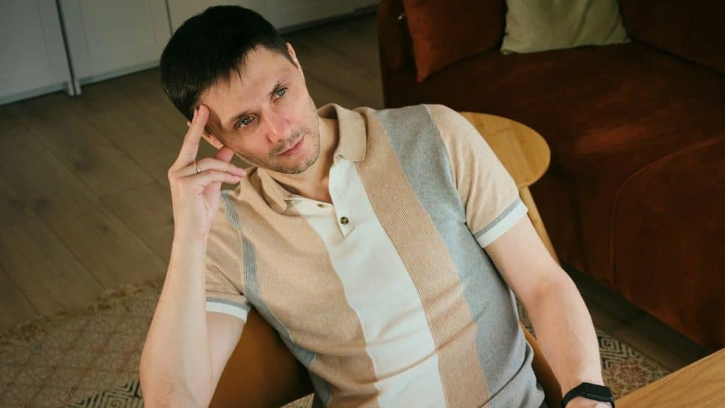 A man in a striped polo shirt is sitting, thoughtfully touching his forehead with one hand.