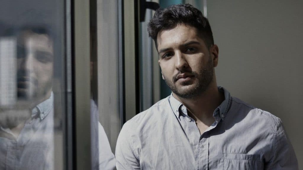 A man with dark hair and a beard is standing by a window, looking directly ahead.