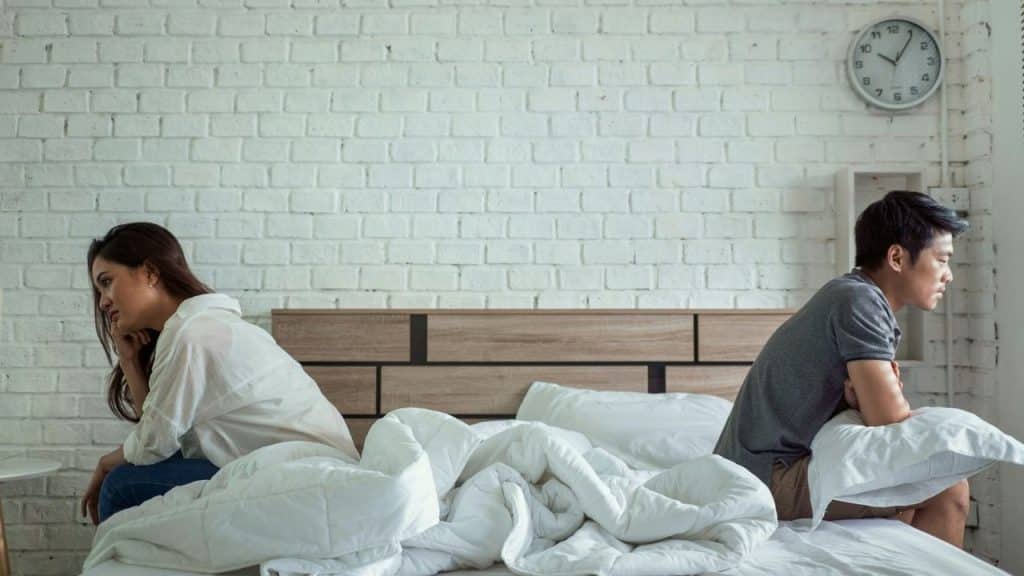 A man and woman sit back-to-back on a bed, looking upset and separate.