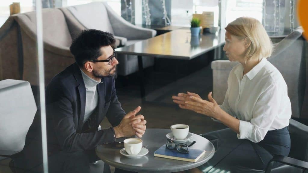 A man and woman sit at a table in a cafe, talking and gesturing.