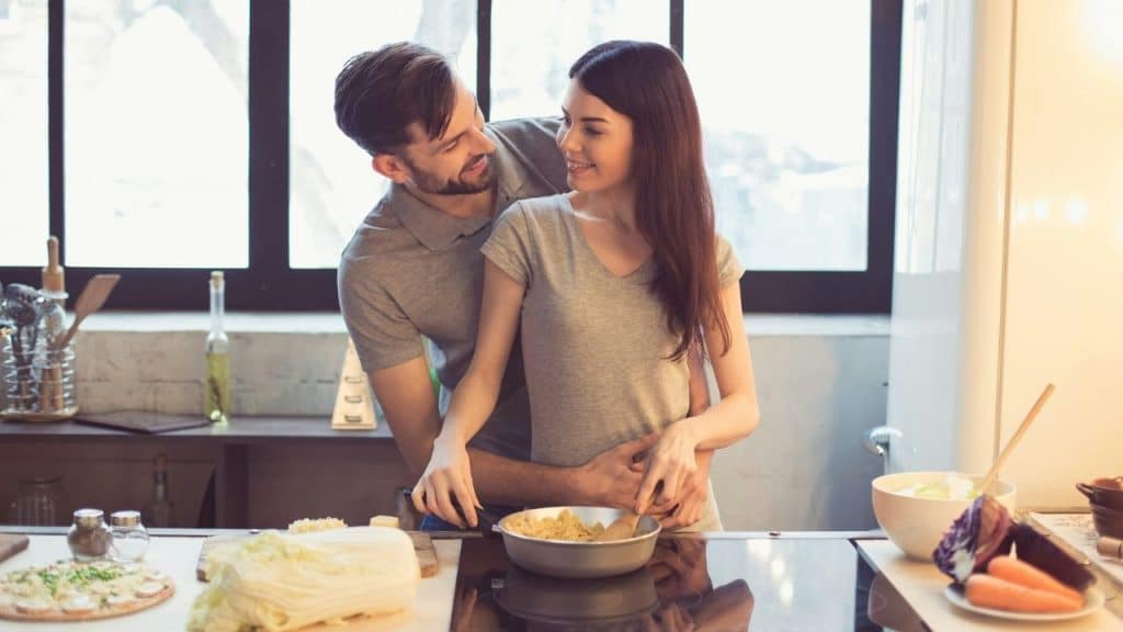 A man hugs a woman from behind as they cook together in a kitchen.