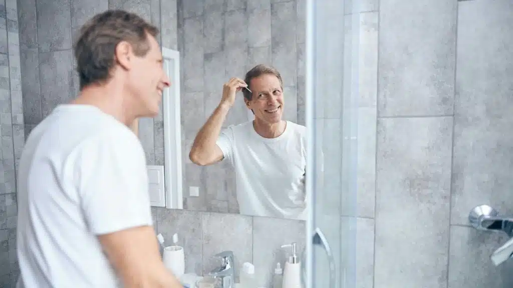 Smiling man in white shirt combing his hair, reflected in a bathroom mirror.