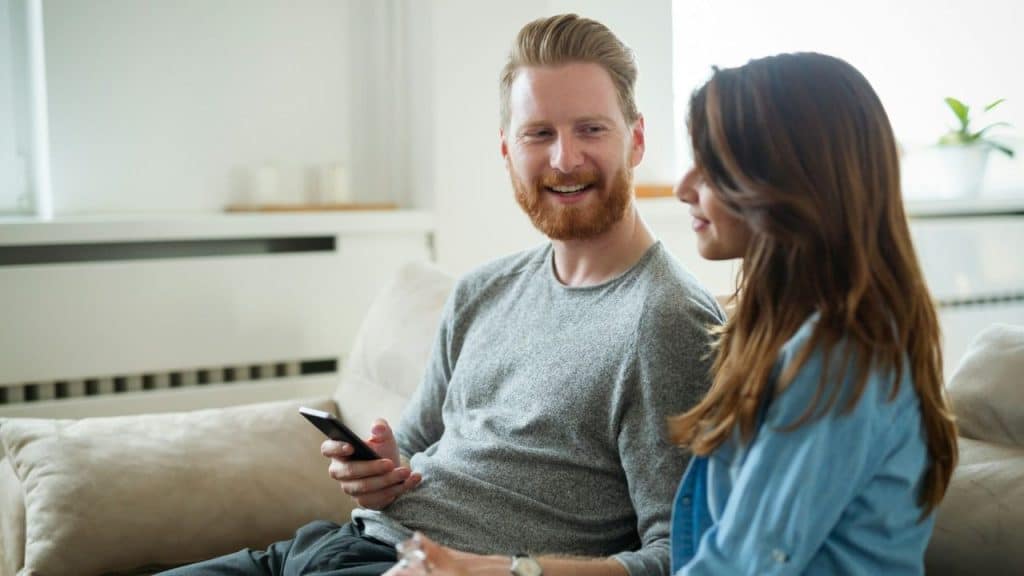 Smiling man holding a phone sits on a couch with a woman.