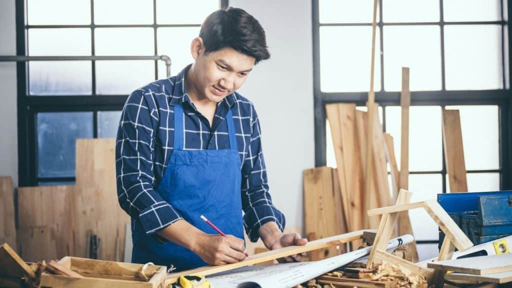 Man in a blue apron drawing on a piece of wood in a workshop.