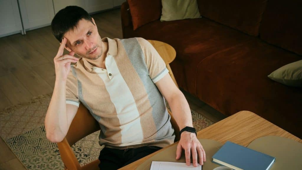 Man sits at a table, looking up and resting his hand on his head.