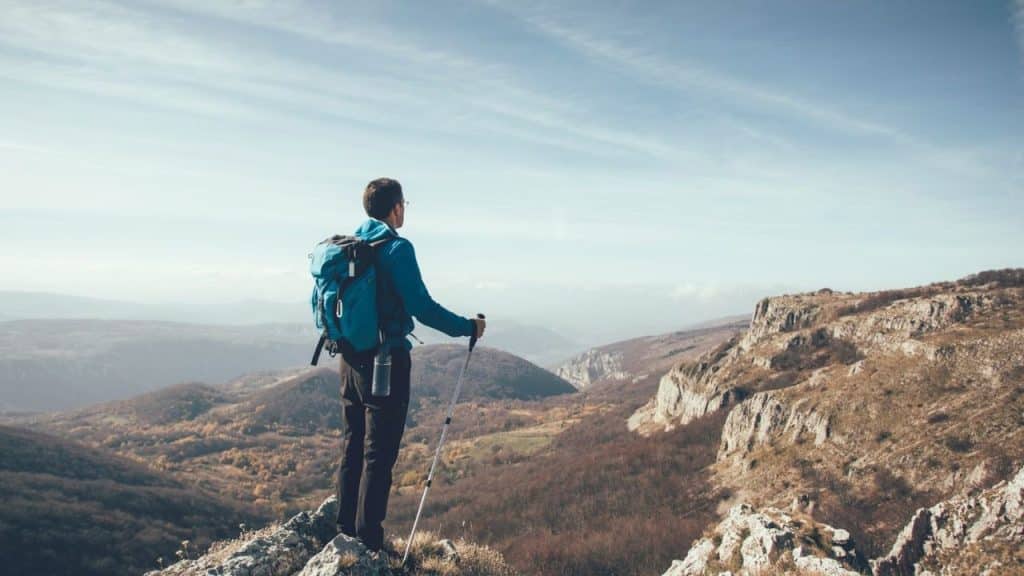 Hiker with a blue backpack and trekking poles standing on a mountain peak.