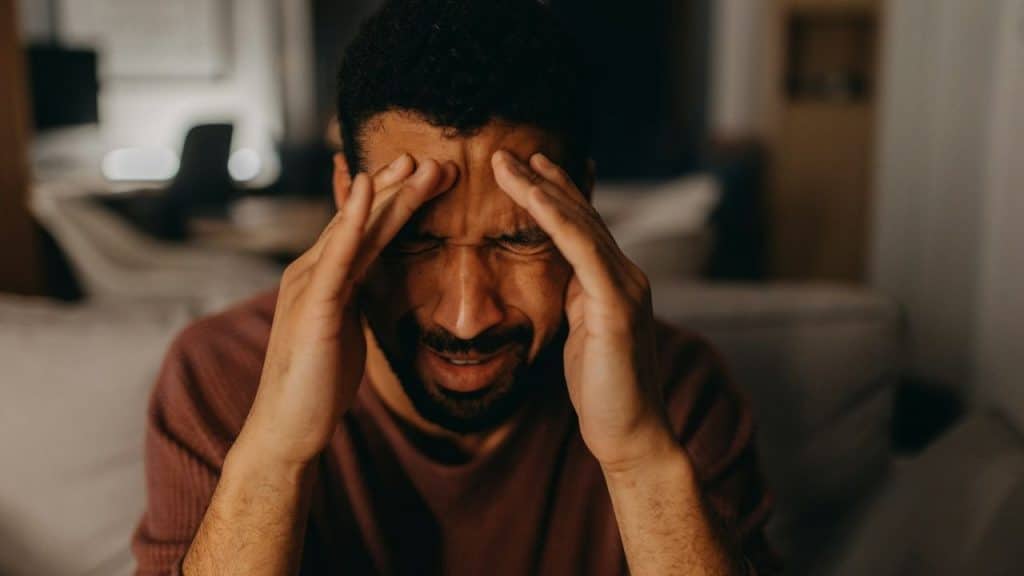 A man grimaces, holding both hands to his forehead in a dimly lit room.