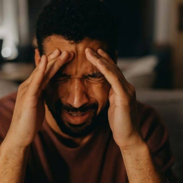 A man grimaces, holding both hands to his forehead in a dimly lit room.
