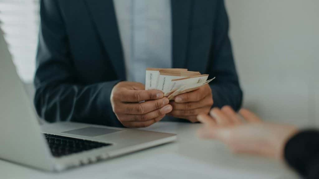 A person in a suit counts cash across a desk from an outstretched hand.