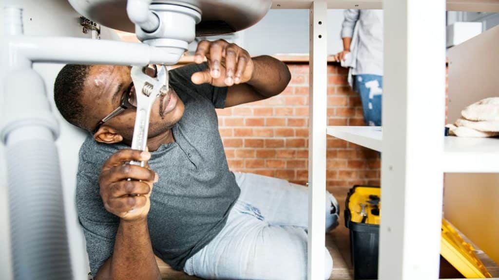 A man fixes a pipe under a sink with a wrench in a brick-walled room.