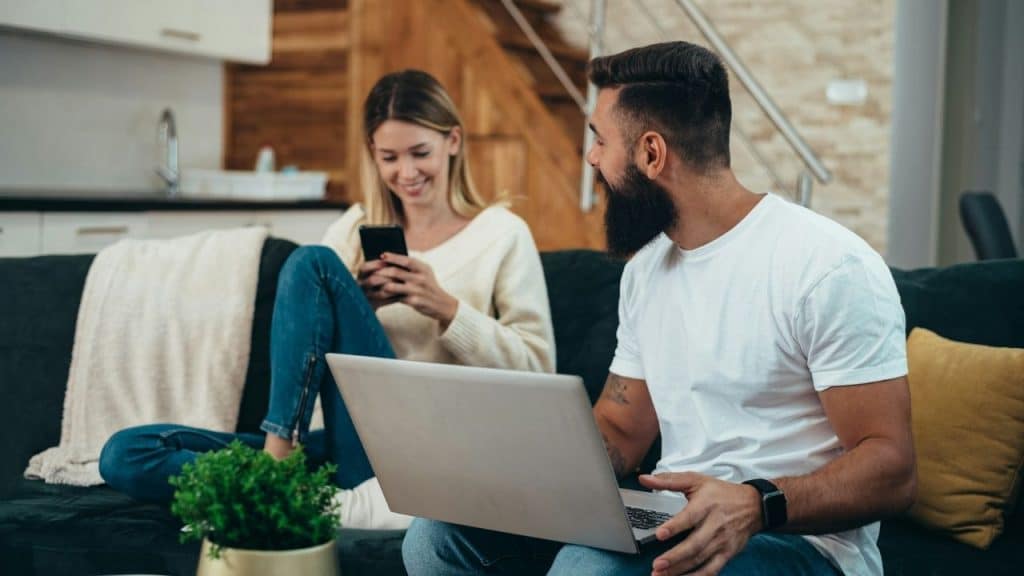 A man with a laptop looks at a woman using a phone on a couch.