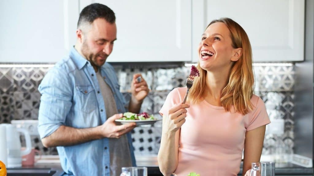 A woman laughs, holding a fork with food, next to a man with a plate.