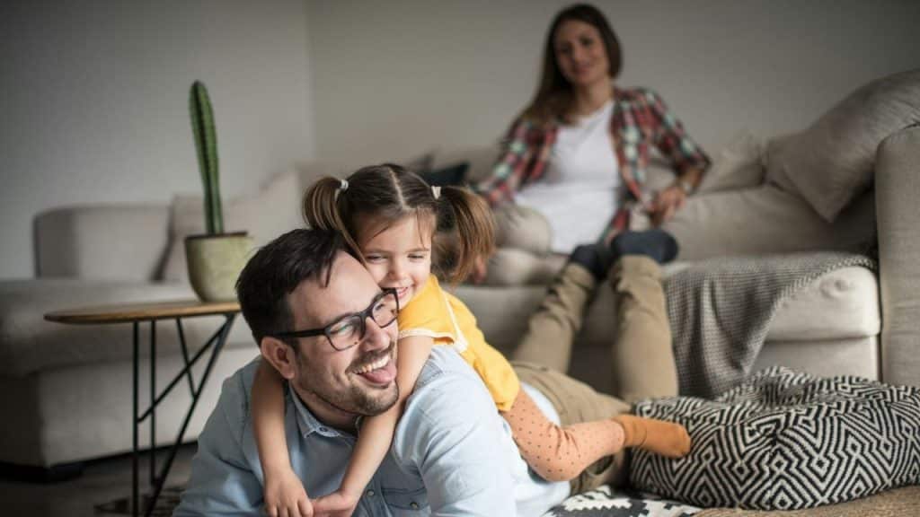 A girl hugs her smiling father on the floor with a woman in the background.