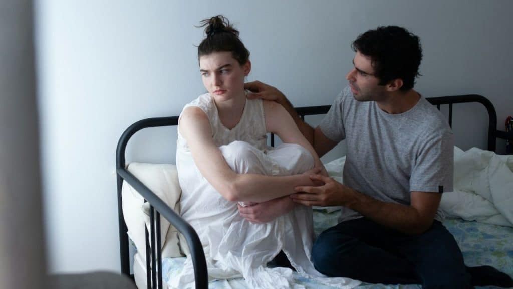 A man comforts a distressed woman sitting on a metal-framed bed.