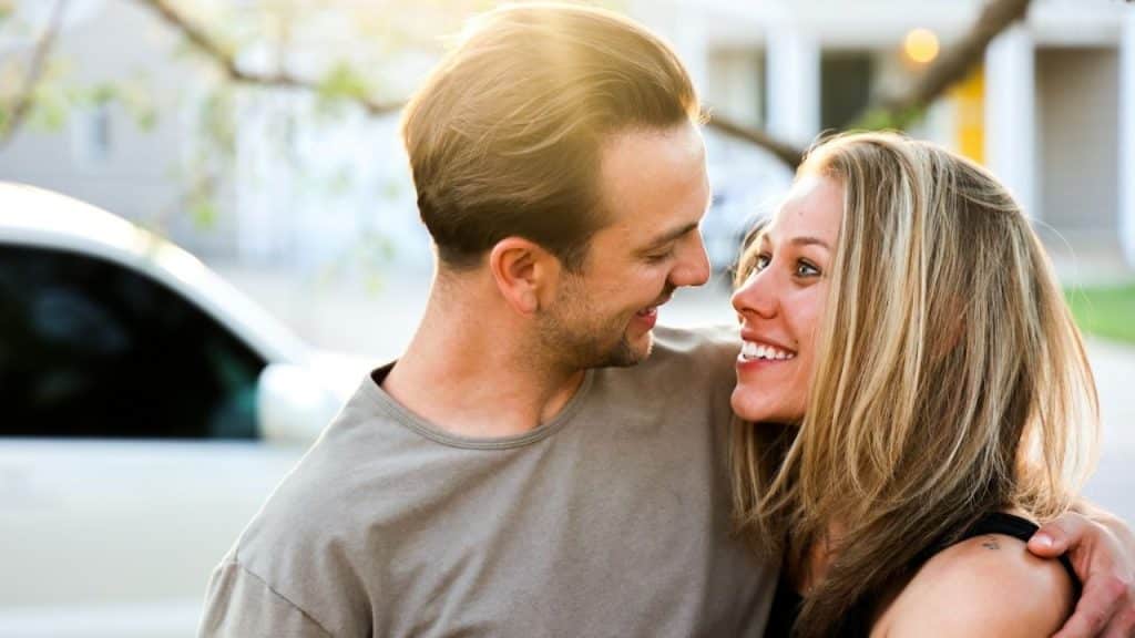 Happy couple embracing and smiling at each other outdoors with a car in the background.