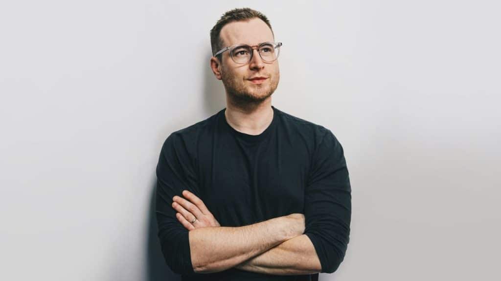 Man wearing glasses and a black shirt, standing with his arms crossed against a white wall.