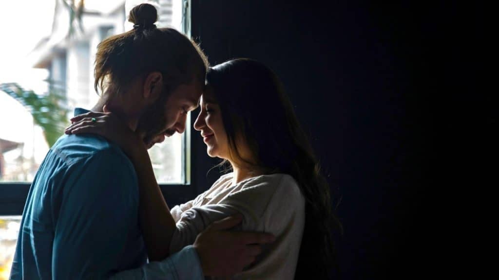 Couple embracing tenderly with foreheads touching in a dimly lit room near a window.
