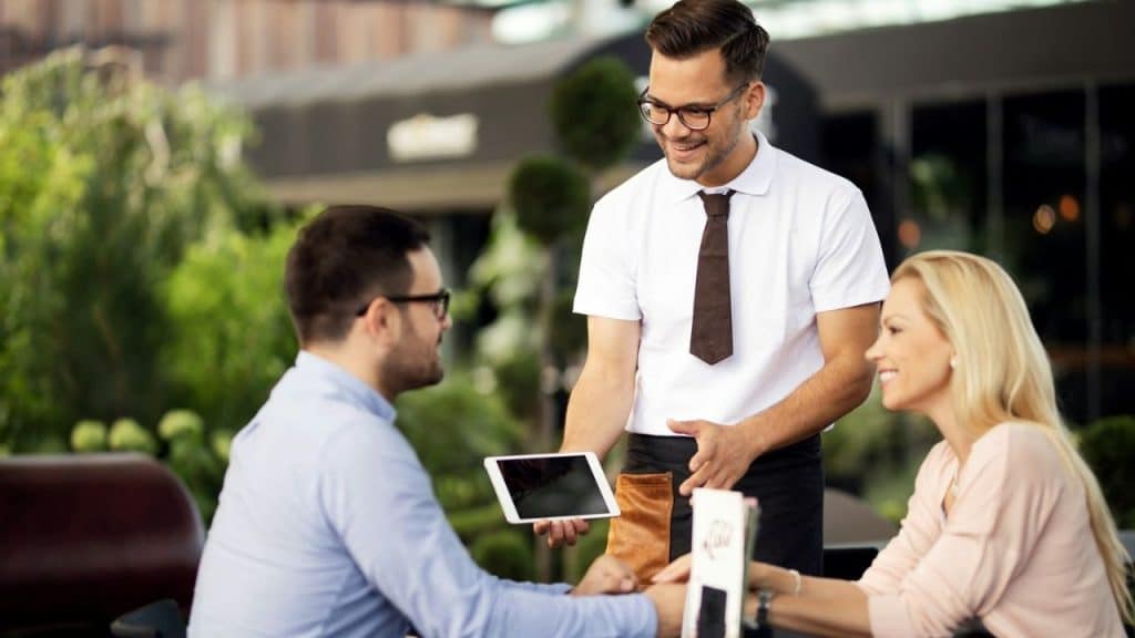 Smiling waiter holding a tablet while taking an order from a couple at an outdoor restaurant.