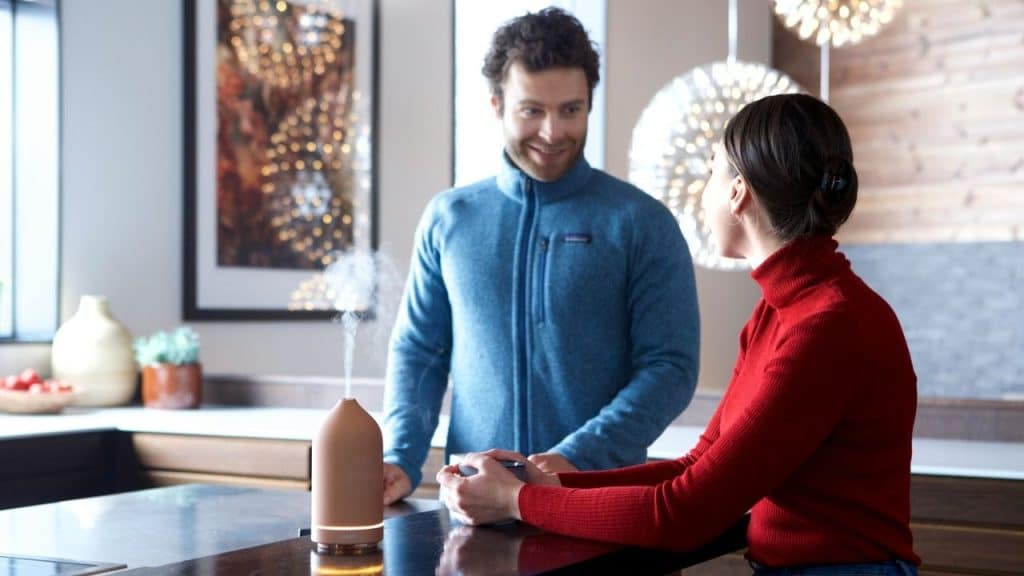 Man in a blue jacket talking to a woman in a red turtleneck across a kitchen counter with a diffuser.