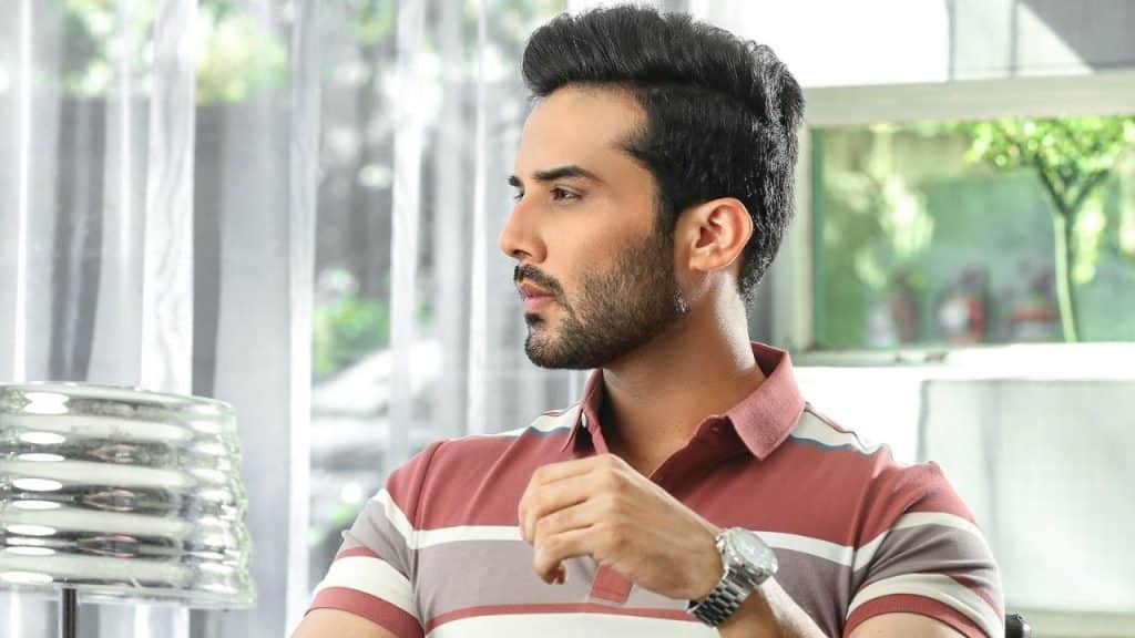 Man in a striped polo shirt and watch looking thoughtfully to his left indoors.