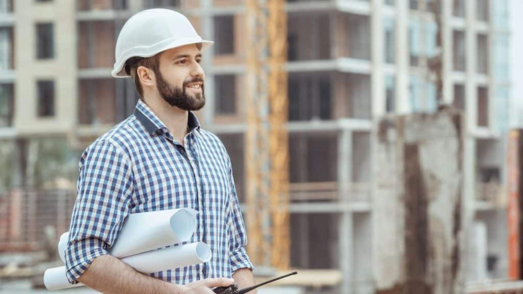 Smiling bearded man in a hard hat holding rolled-up blueprints at a construction site.