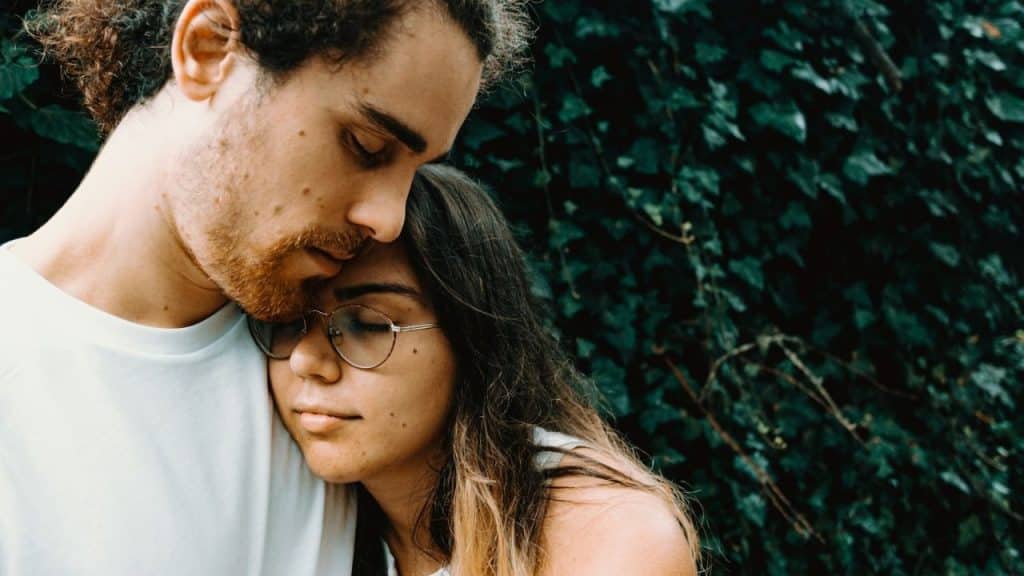 Man holding a woman wearing glasses, with her head resting on his chest, against a dark leafy background.
