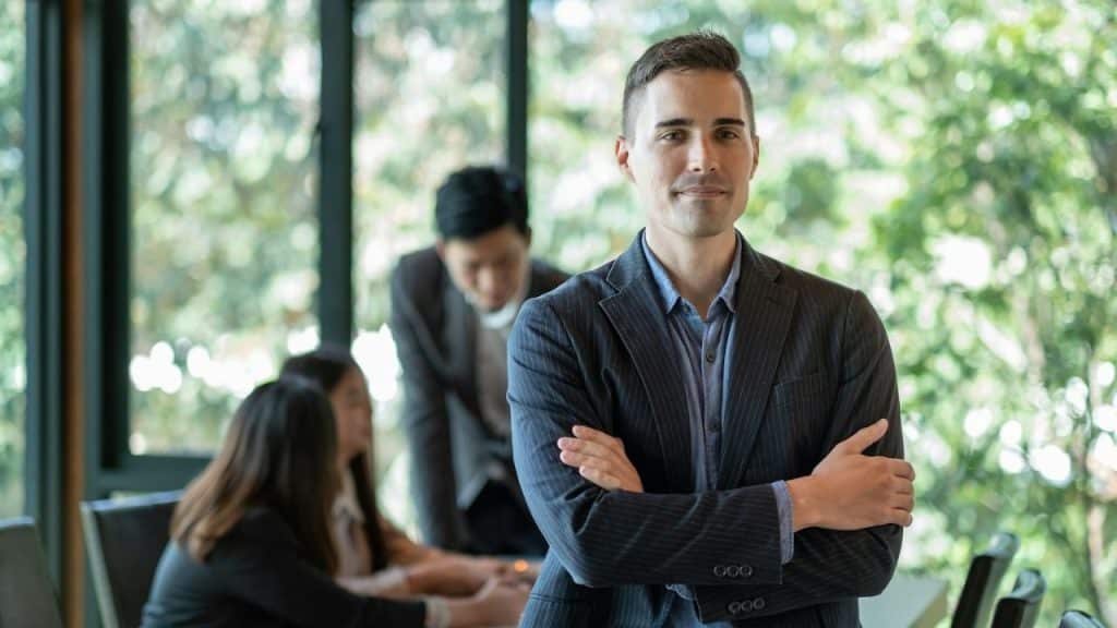 Man in a suit standing with crossed arms in an office setting with people blurred in the background.