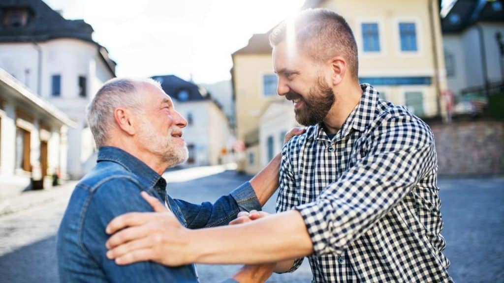 Two men smiling at each other and reaching for a hug on a sunny European street.