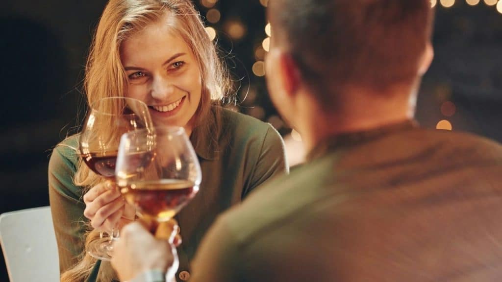 Smiling woman clinking wine glasses with a man in a dark, warm-lit setting.