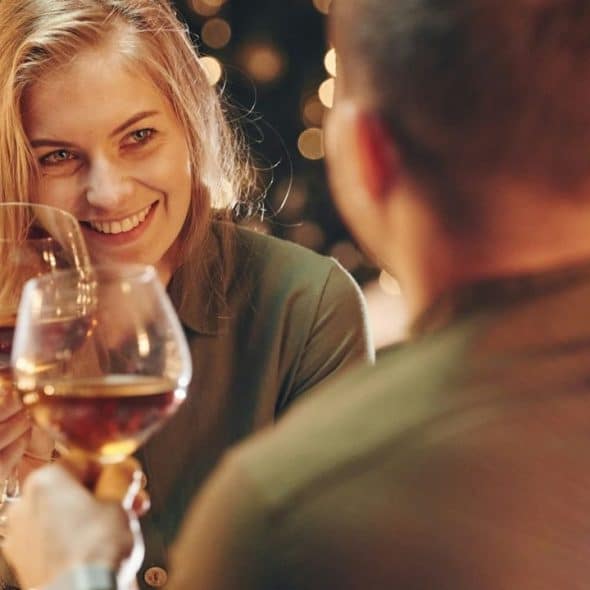 Smiling woman clinking wine glasses with a man in a dark, warm-lit setting.