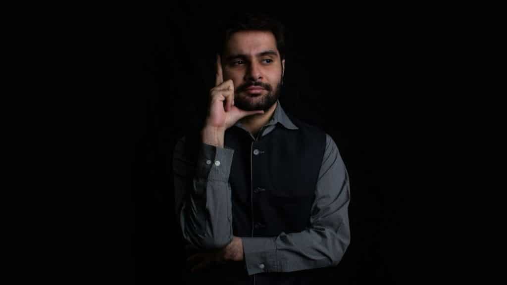 Man in a gray shirt and black vest with his hand on his chin, looking thoughtful against a black background.