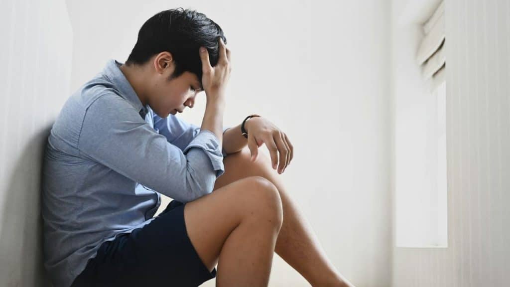 Young man sitting on the floor with his head in his hands, looking distressed indoors.