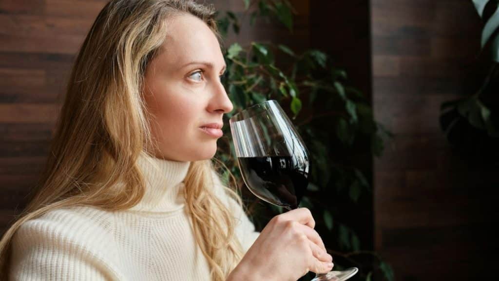 Blonde woman in a white turtleneck smelling red wine in a glass indoors.