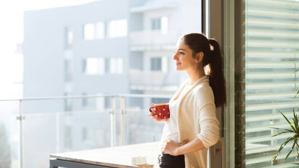 Woman holding a red polka-dot mug, looking out from a balcony on a sunny day.