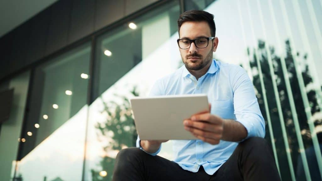 Man in a blue shirt and glasses sitting outdoors, intently looking at a tablet.