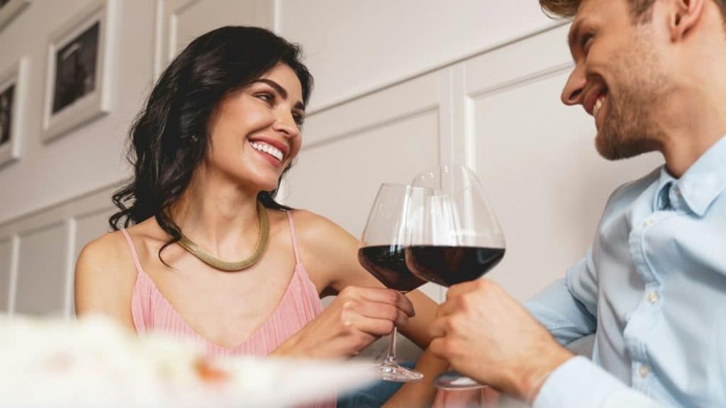 Smiling woman in a pink top clinking red wine glasses with a man.