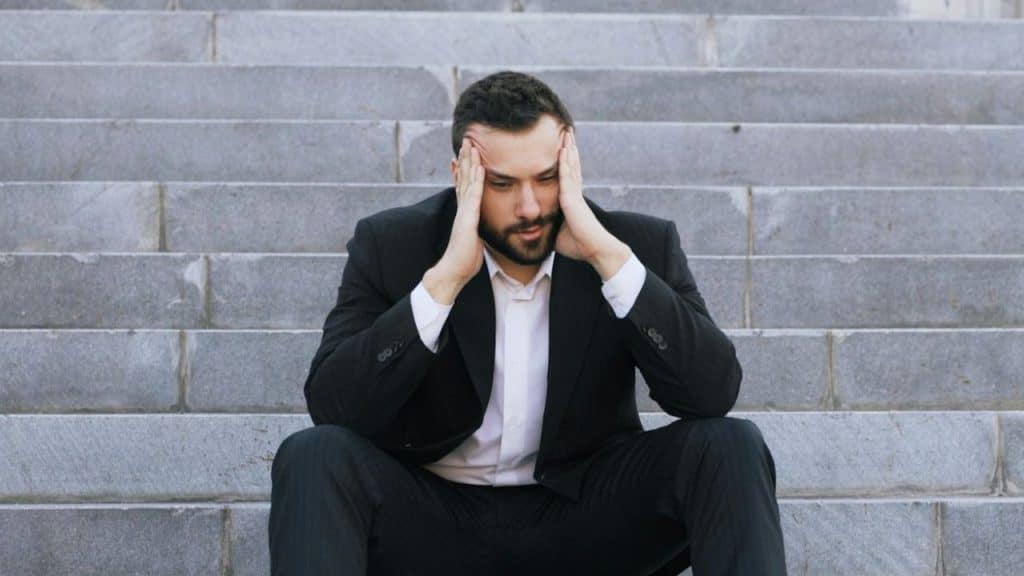Man in a dark suit sitting on concrete steps with his hands held to his temples.