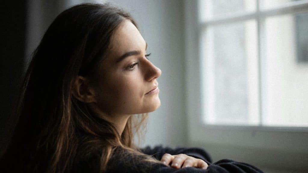Woman with long brown hair looking out a bright window with a thoughtful expression.