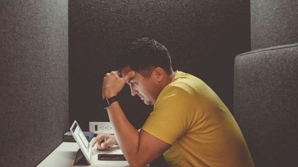 Man in a yellow shirt with his head in his hand, looking stressed at a laptop in a private booth.