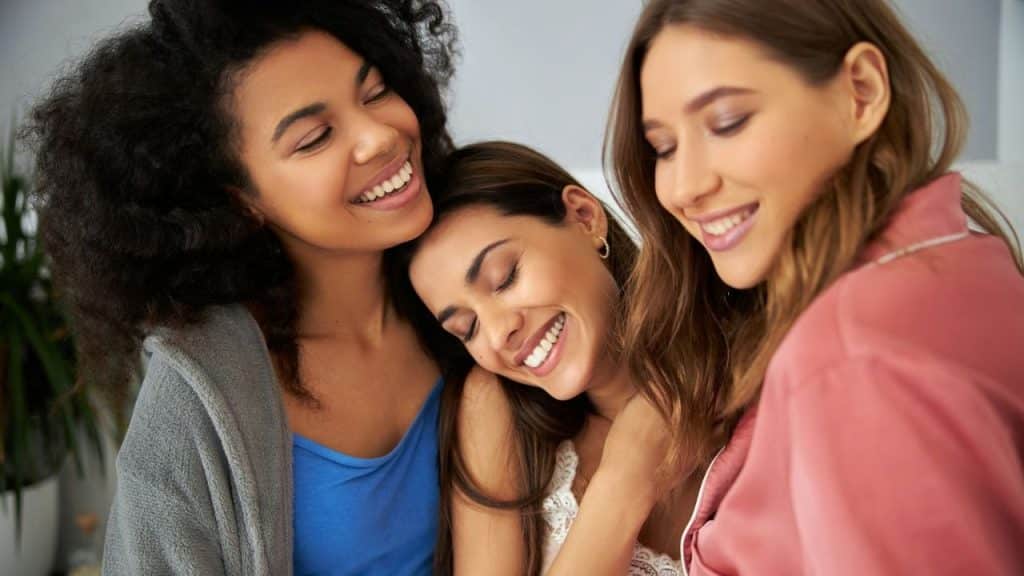 Three young women happily embracing and smiling together in a close-up indoor shot.