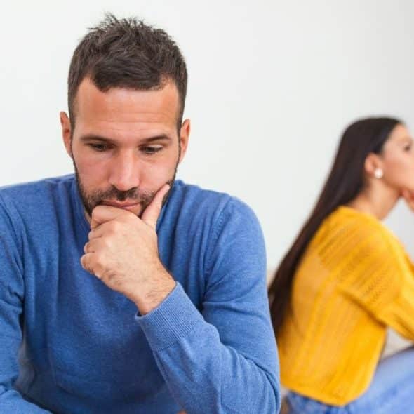 A sad man sits in the foreground, contemplating, with a woman blurred in the background.